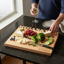 A person arranges cheese, grapes, blueberries, cucumbers, and green peppers on a large (16” x 12.5”) Words With Boards Seriously Kosher Personalized Cutting Board by Words with Boards, LLC while holding a glass of white wine.