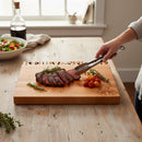 Someone uses tongs to serve steak garnished with rosemary and cherry tomatoes on a Words With Boards Wedding Bells Personalized Cutting Board 12.5" x 16" by Words with Boards, LLC—an ideal wedding gift for any sunlit kitchen table.
