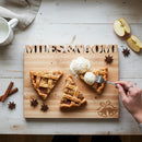 The Words With Boards Wedding Bells Personalized Cutting Board (12.5" x 16") by Words with Boards, LLC, engraved with MILES & NAOMI, displays apple pie slices, vanilla ice cream, and spices—ideal for a memorable wedding gift.