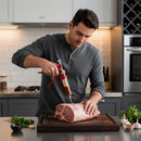 A man in a gray shirt uses the DDR BBQ Gourmet Pistol Grip Meat Marinade Injector with Case by DDR BBQ Supply to season a large raw pork roast on a kitchen counter, surrounded by fresh herbs, garlic, and spices.