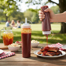 A hand squeezes DDR BBQ Supply Plastic Squeeze Bottles for Sauces – 24 oz onto grilled ribs on a plate, surrounded by sauce bottles, sides, and a pitcher of iced tea on an outdoor picnic table with people in the background.