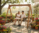 A couple enjoys drinks and smiles at each other on the Kanyon Living Poly Lumber 4′ Adirondack Swing Deluxe in Antique Mahogany and Black, surrounded by colorful potted flowers on a sunny stone patio.
