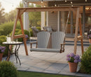 The Kanyon Living Poly Lumber 4′ Adirondack Swing in Light Grey and Weathered Wood hangs on a stone patio. Nearby, a small round table with refreshments and potted purple flowers enhance the relaxing outdoor space.