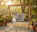 The Kanyon Living Poly Lumber 4′ Adirondack Swing in Light Grey and Weathered Wood sits on a stone patio with cushions, a knit blanket, sunhat, potted plants, and flowers amid trees and soft sunlight.