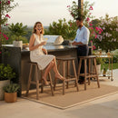 A woman in a light dress sits laughing with a man by an outdoor bar on Kanyon Living Poly Lumber Saddle Stool Bar Height Deluxe Brazilian Walnut and Weathered Wood barstools, surrounded by plants and drinks, enjoying a relaxed, sunny atmosphere.