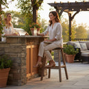 Two women enjoy drinks at an outdoor stone bar, seated on Kanyon Living Poly Lumber Saddle Stools Bar Height Deluxe in Brazilian Walnut and Weathered Wood, surrounded by patio furniture and plants in the soft evening light for a relaxed atmosphere.