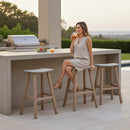 A woman in a light dress sits on a Kanyon Living Poly Lumber Saddle Stool Bar Height Light Grey and Weathered Wood at an outdoor kitchen counter, smiling with a drink. A grill, fruit plate, and two empty barstools are beside her in the bright, modern setting.