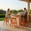 A woman smiles and holds a drink while sitting on a Kanyon Living Poly Lumber Saddle Stool Bar Height in Dark Wood and Orange, as a man grills at an outdoor kitchen with greenery and food on the counter in a bright, relaxed setting.