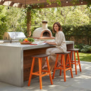 A woman sits on a Kanyon Living Poly Lumber Saddle Stool Bar Height in Dark Wood and Orange at an outdoor kitchen island, smiling amid fresh vegetables, a pizza oven, greenery, and string lights.