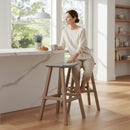 A woman in light, casual clothes sits on a Kanyon Living Poly Lumber Saddle Stool Bar Height in Light Grey and Weathered Wood at a bright kitchen counter with coffee, croissant, and lavender. Sunlight fills the modern, minimalist space.