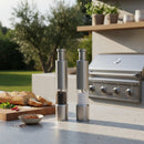 Two DDR BBQ Supply Thumb Grinders from DDR BBQ Supply sit on an outdoor kitchen counter, surrounded by sliced bread, spices, and a gas grill. Perfect for effortless one-handed seasoning. Trees can be seen in the garden background.