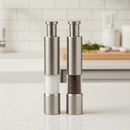 Two DDR BBQ Supply Thumb Grinders by DDR BBQ Supply sit on a white countertop, ideal for one-handed seasoning. Their stainless steel, push-button design and modern setting make them must-have BBQ accessories.
