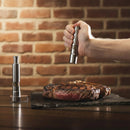 A hand seasons a cooked steak on a slate board using DDR BBQ Supply Thumb Grinders by DDR BBQ Supply, with another stylish grinder upright nearby. A brick wall background highlights these must-have BBQ accessories.