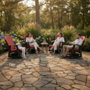 Three people sit and chat on Kanyon Living Adirondack Poly Glider Swivel Chairs in red and black, enjoying a garden patio filled with blooming hydrangeas and trees. Sunlight streams through as drinks and a book rest on small tables between them.
