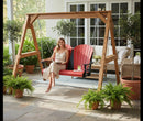 A woman in a light dress smiles while sitting on a Kanyon Living Poly Lumber 4′ Adirondack Swing in red and black, enjoying a drink on a patio with potted plants, outdoor furniture, and large windows behind her.