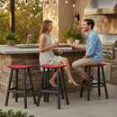 A woman in a light dress and a man in a blue shirt relax on Kanyon Living Poly Lumber Saddle Stool Bar Height Red and Black seats at an outdoor kitchen, smiling with drinks, surrounded by greenery and patio lights.