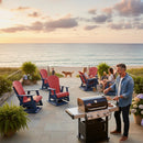 A man grills on a patio overlooking the ocean at sunset while three people relax in Kanyon Living Adirondack Poly Glider Swivel Chairs in red & blue, with a dog strolling nearby and potted plants adding eco-friendly style.