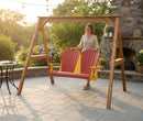 A woman stands by the Kanyon Living Poly Lumber 4′ Adirondack Swing in red and yellow on a stone patio with greenery, flowers, string lights, outdoor furniture, a stone fireplace, and a table nearby.
