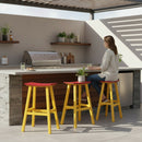 A woman sits at a modern outdoor kitchen counter furnished with Kanyon Living Poly Lumber Saddle Stool Bar Height Red and Yellow by Kanyon Living, surrounded by potted plants and a built-in grill under a partially shaded pergola.
