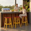 A man and woman smile and chat at an outdoor bar featuring a Kanyon Living Poly Lumber Saddle Stool Bar Height in red and yellow. The sturdy, weather-resistant stool by Kanyon Living adds style to the sunlit, inviting space filled with greenery and glasses.