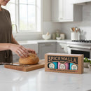 In a bright kitchen, someone slices bread on a wooden board while the Spicewalla Salt Collection 3 Pack by Spicewalla, featuring three specialty finishing salts, sits on the countertop in the foreground.