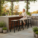 A woman sits at an outdoor bar on three Kanyon Living Poly Lumber Saddle Stool Bar Height stools in Seashell and Black, surrounded by potted plants, a grill, and bottles, with greenery and trees in the background.