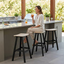 A woman sits smiling on a Kanyon Living Poly Lumber Saddle Stool Bar Height in Seashell and Black at an outdoor kitchen counter, holding orange juice. The counter has potted plants, drinks, and two empty Kanyon Living barstools beside her.