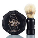 DDR BBQ Supply Immaculate Beard shave soap puck in black paper wrapping next to a wooden handle shaving brush
