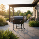 A Smokin Brothers “30” Premier Plus Pellet Grill stands on a paver patio with plants and outdoor furniture under a pergola at sunset. This American-made black grill features wheels for easy movement.