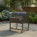 A Smokin Brothers “36” Premier Plus Pellet Grill by Smokin Brothers sits on a stone patio surrounded by green bushes, hydrangeas, and a wooden fence, wood-fired smoke rising from its precision digital controls.