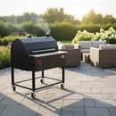 The Smokin Brothers “36” Premier Plus Pellet Grill stands on a stone patio amid wicker furniture and lush greenery, smoke rising from its chimney as it fills the air with wood-fired flavor on a sunny day.
