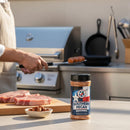 A bottle of Texas Pepper Jelly Southern Pecan Seasoning sits on an outdoor counter beside pecans and pork chops, while someone grills sausages nearby, filling the air with smoky aroma from the grill.