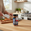 A jar of Texas Pepper Jelly Southern Pecan Seasoning rests on a sunlit kitchen counter as someone uses its smoky, savory spices to prepare salmon amid white cabinets.