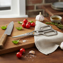A kitchen scene featuring Spaceman Knives Kitchen Shears and a knife on a napkin with cherry tomatoes, basil, garlic, rosemary, salt, and olive oil atop a wooden cutting board. Brick wall and window in the background.