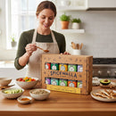 A woman in an apron smiles in her kitchen as she measures out Spicewalla Signature Blends 10-Pack from Spicewalla, surrounded by bowls of fresh vegetables, grains, and pita bread on the counter.