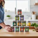 A woman stands at a kitchen counter beside a neatly arranged pyramid of ten Spicewalla Signature Blends tins. Fresh herbs and lemons accent the bright, modern kitchen—Spicewalla’s 10-Pack is the perfect seasoning gift set for any cook.