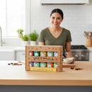 A woman smiles in a modern kitchen, standing behind a box of Spicewalla Signature Blends 10-Pack by Spicewalla on the counter, surrounded by global spices ready to inspire creative cooking.