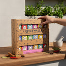 A hand reaches for the Spicewalla Kitchen Essentials 18-Pack Collection on a wooden countertop surrounded by herbs, a mortar and pestle, cinnamon sticks, and dried chili peppers—must-have essentials for any cook.