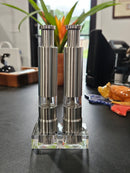 The DDR BBQ Supply Thumb Grinders by DDR BBQ Supply feature stainless steel and clear mid-sections, standing on a transparent base. Ideal as BBQ accessories, these one-handed grinders sit on a dark counter with other items and a plant behind them.