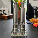 Two DDR BBQ Supply Thumb Grinders by DDR BBQ Supply, featuring clear sections, stand on a glass base atop a dark countertop with kitchen items and a colorful ceramic ornament in the background.