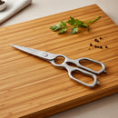 Spaceman Knives Kitchen Shears, featuring stainless steel blades, rest on a wooden cutting board. Ideal for spatchcocking poultry, this multi-functional tool is neatly arranged beside parsley and black peppercorns.
