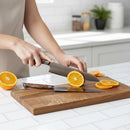 In a bright kitchen, a person slices an orange with a knife from the Steelport Knife Co. 3-Piece Essential Set on a wooden cutting board. Another Steelport Knife Co. knife and several more orange slices are also on the cutting board.