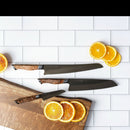 Three Steelport Knife Co. 3-Piece Essential Set kitchen knives with wooden handles are displayed on a white tile surface, surrounded by sliced oranges, with some orange slices resting on a wooden cutting board in the lower left corner.