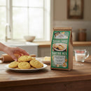 A hand reaches for a homemade cookie from a plate on a wooden counter. Beside the plate is a Fowler's Mill Sugar Cookie Baking Mix bag, with various baking ingredients and utensils in the background.