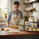 Wearing an apron, a woman rolls out dough on a floured counter beside homemade cookies and a bag of Fowler's Mill Sugar Cookie Baking Mix, creating a cozy kitchen scene.