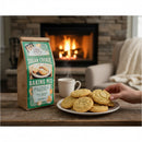 A hand reaches for a sugar cookie on a plate by a cozy fireplace. Next to it are a steaming mug and a Fowler's Mill Sugar Cookie Baking Mix bag, making the scene warm and inviting.
