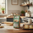 A bag of Fowler's Mill Sugar Cookie Baking Mix sits on a wooden counter beside homemade cookies, a rolling pin, and baking sheets, with potted herbs, jars, and utensils in the kitchen background.
