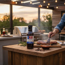 DDR BBQ Supply Sweet Bourbon Glaze bottle on a wooden table with a person brushing sauce onto grilled ribs outdoors.