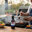 DDR BBQ Supply Sweet Bourbon Glaze bottle on a table while a person brushes sauce onto ribs on a backyard grill.