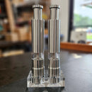 Two DDR BBQ Supply Thumb Grinders by DDR BBQ Supply stand upright on a clear rectangular base, allowing one-handed seasoning with their sleek metallic design. Shelves and a window appear blurred in the background.